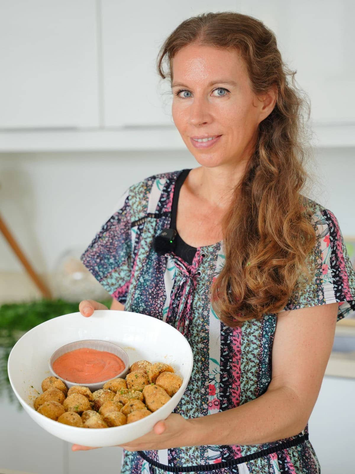 Michaela Vais in a colorful pattern dress smiling and holding a large white bowl filled with crispy potato balls