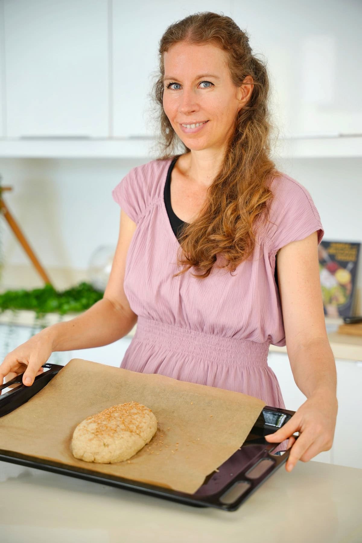 Michaela Vais stands smiling in a kitchen, holding a baking tray with a round loaf of bread