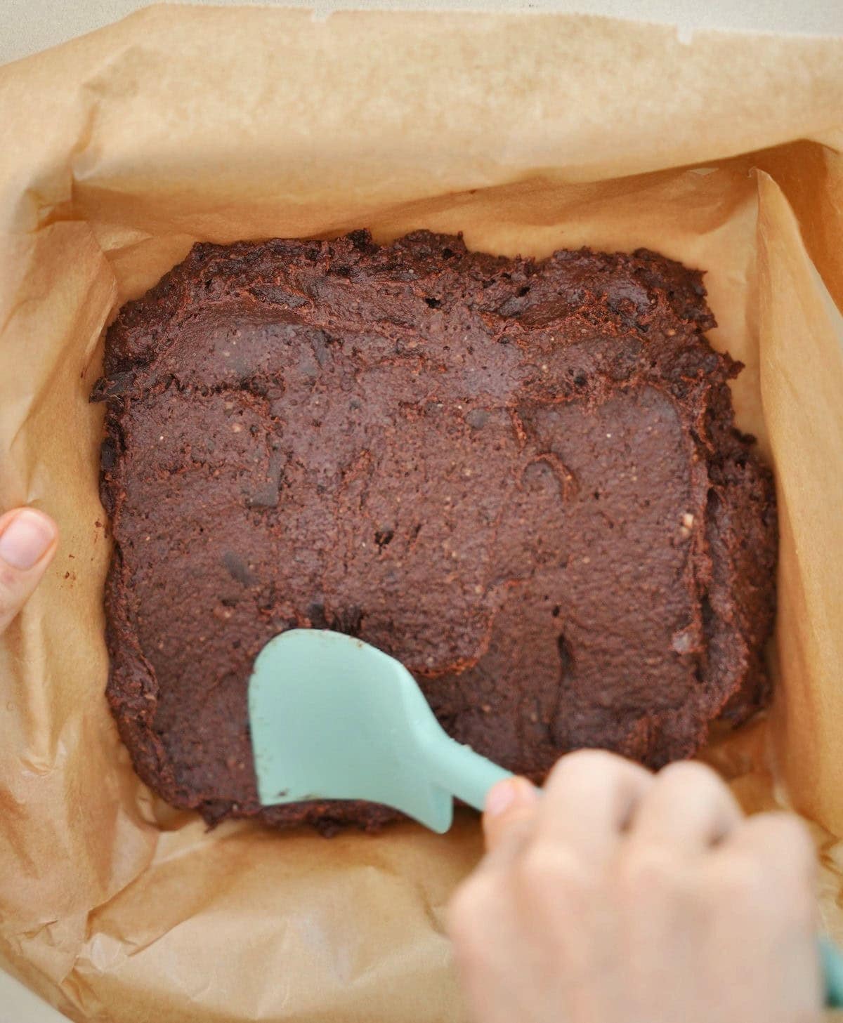 Person spreading chocolate batter in a parchment-lined pan with a teal spatula