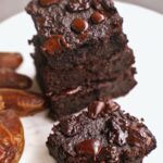 Stack of rich, moist chocolate brownies topped with glossy chocolate chips on a white plate. Nearby are whole dates
