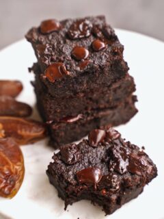 Stack of rich, moist chocolate brownies topped with glossy chocolate chips on a white plate. Nearby are whole dates