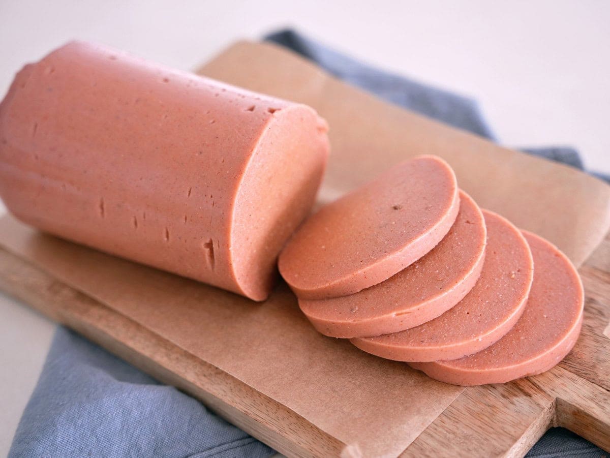 A block of vegan bologna lies on a wooden cutting board partially sliced