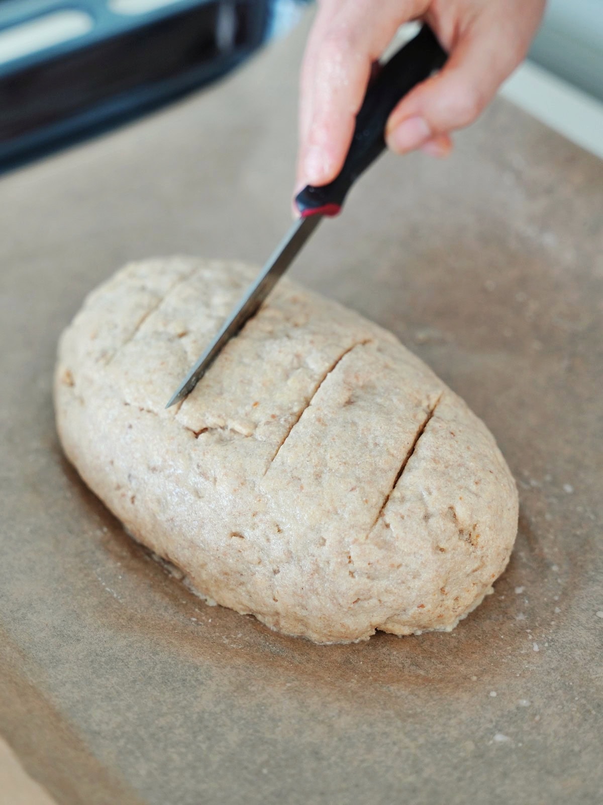 A hand holds a knife, scoring a loaf of dough on parchment paper
