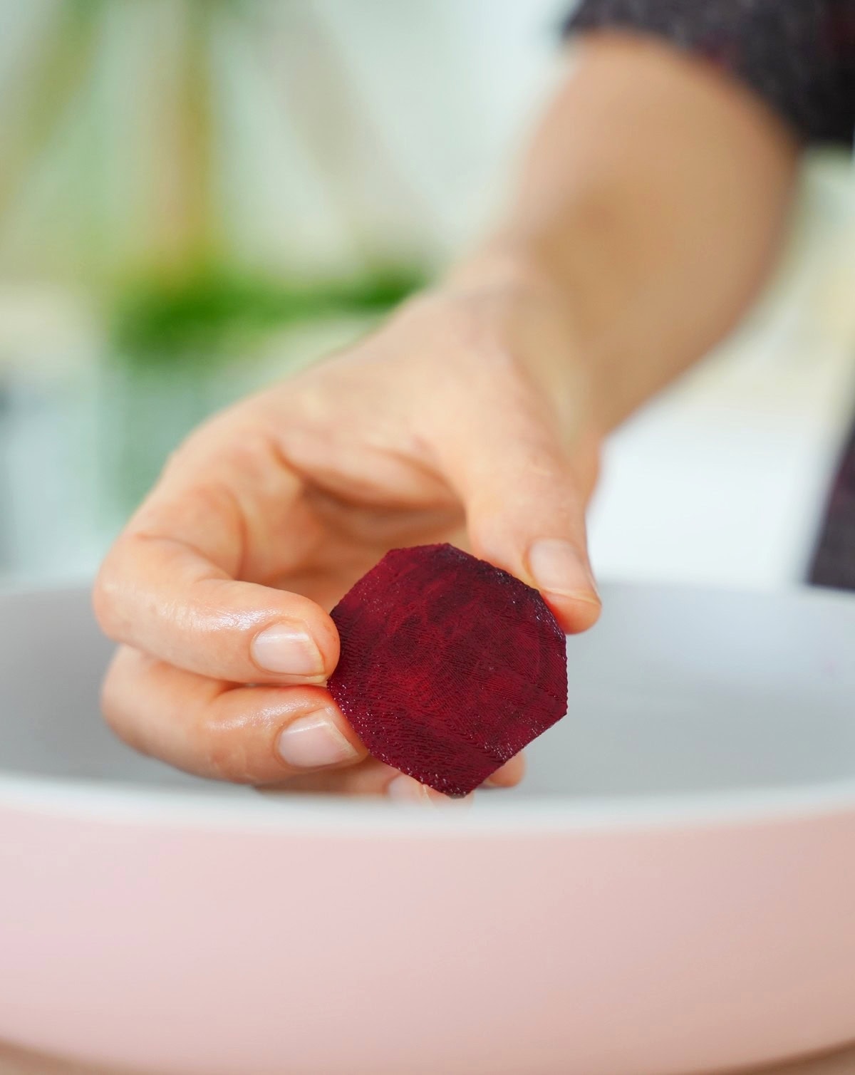 A hand holds a small, round slice of beetroot over a white bowl with a pink rim
