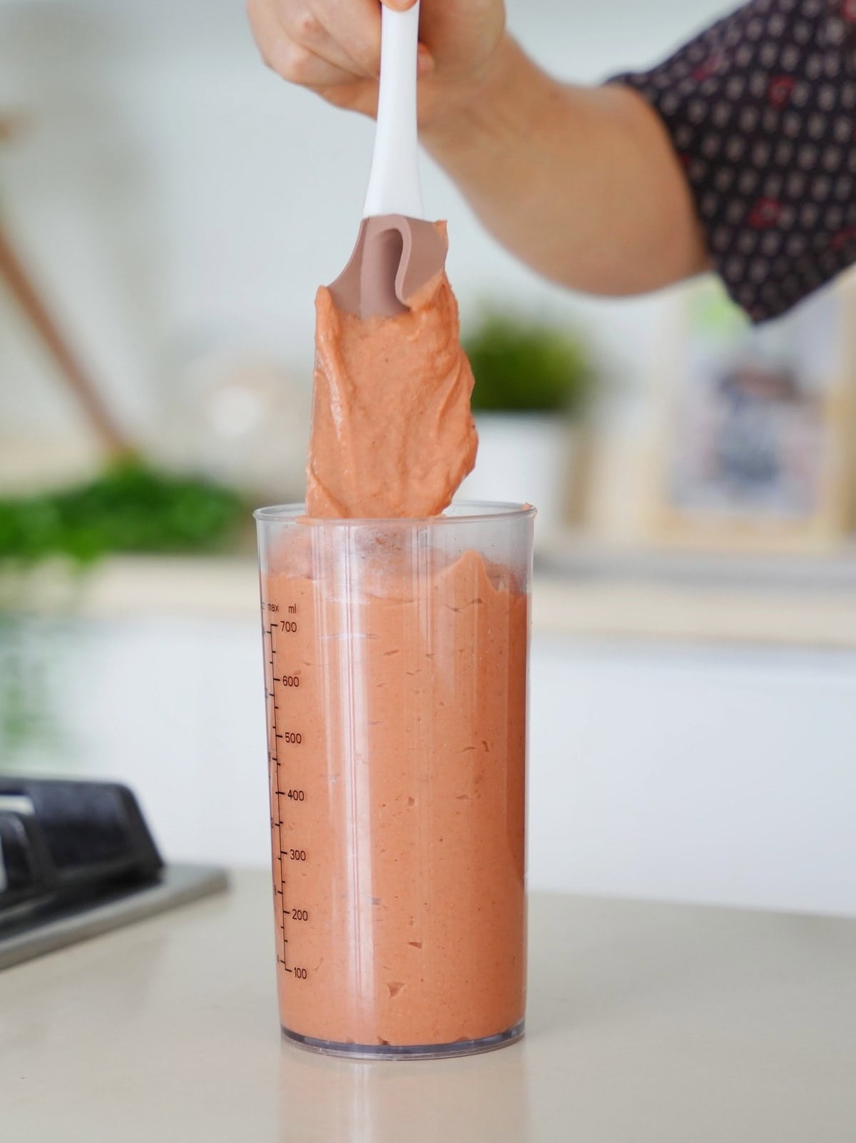 A hand holds a spatula with creamy, light brown dough lifting it from a tall measuring cup filled with the same mixture