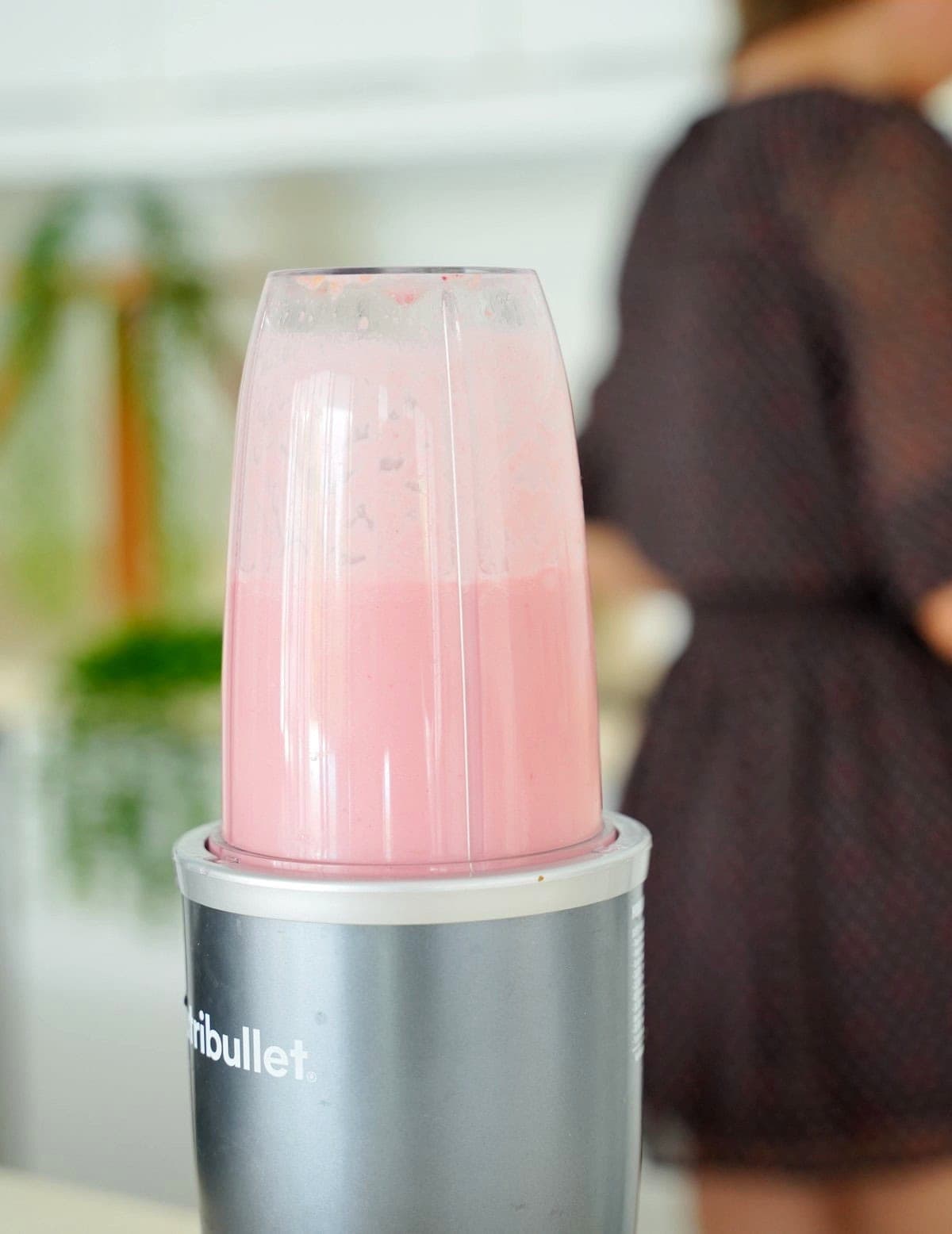 Close-up of a blender filled with pink liquid in a bright kitchen. Blurred woman in dress is in the background