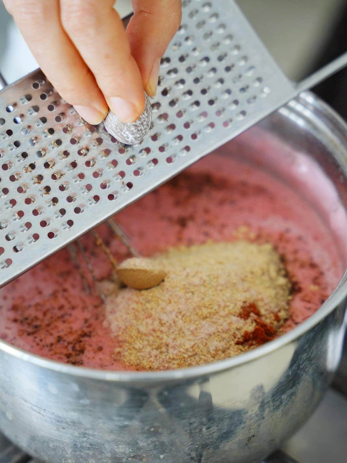 Close-up of a hand grating nutmeg over a pot of pink liquid