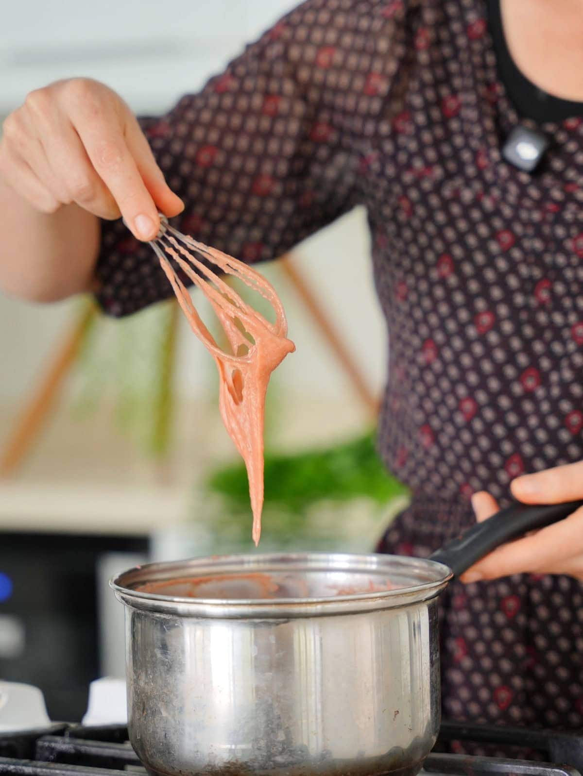 Elavegan in a patterned dress stirs thick, creamy sauce using a whisk over a stainless-steel saucepan on a stovetop