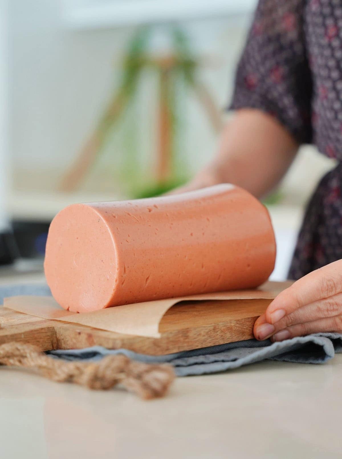 Elavegan places a smooth, round log of vegan bologna on a wooden cutting board lined with paper
