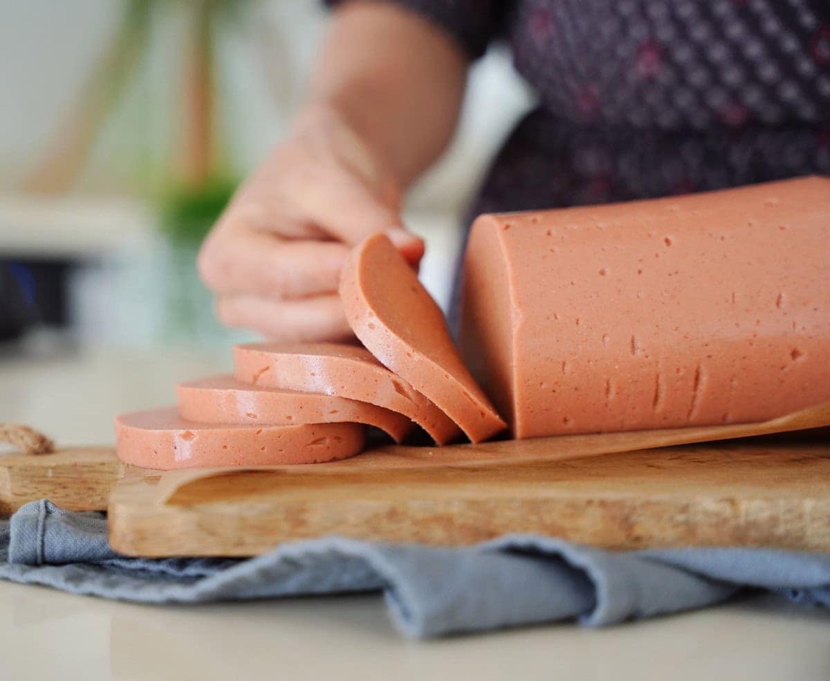 Elavegan slices a log of vegan bologna on a wooden cutting board. Several slices are neatly arranged