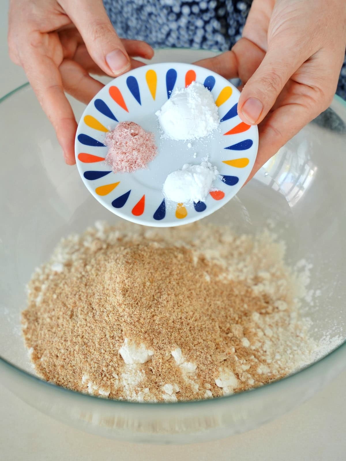 Hands holding a small plate with salt and baking powder, soda, over a mixing bowl