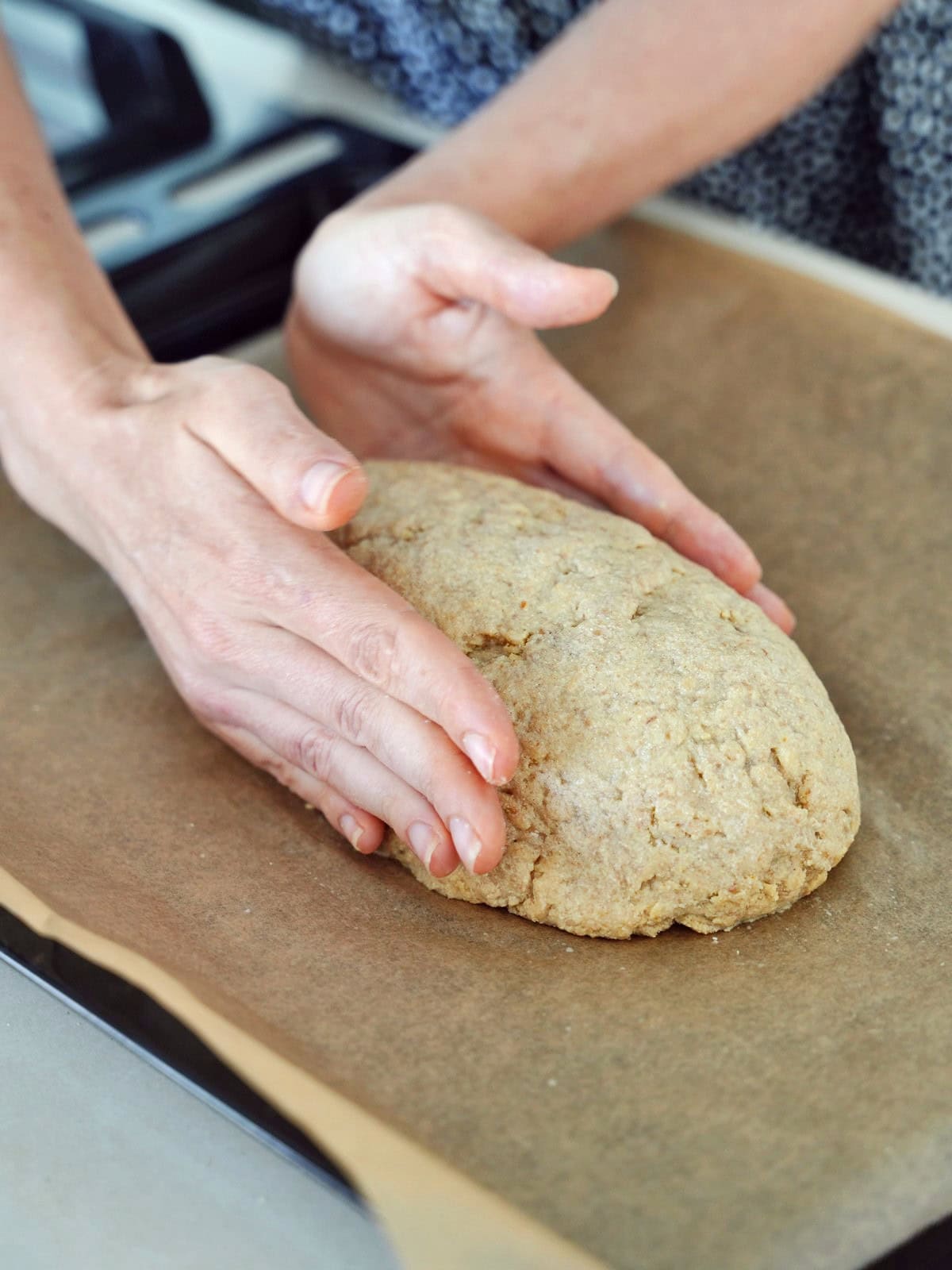 Hands shaping a loaf of dough on parchment paper, resting on a baking tray