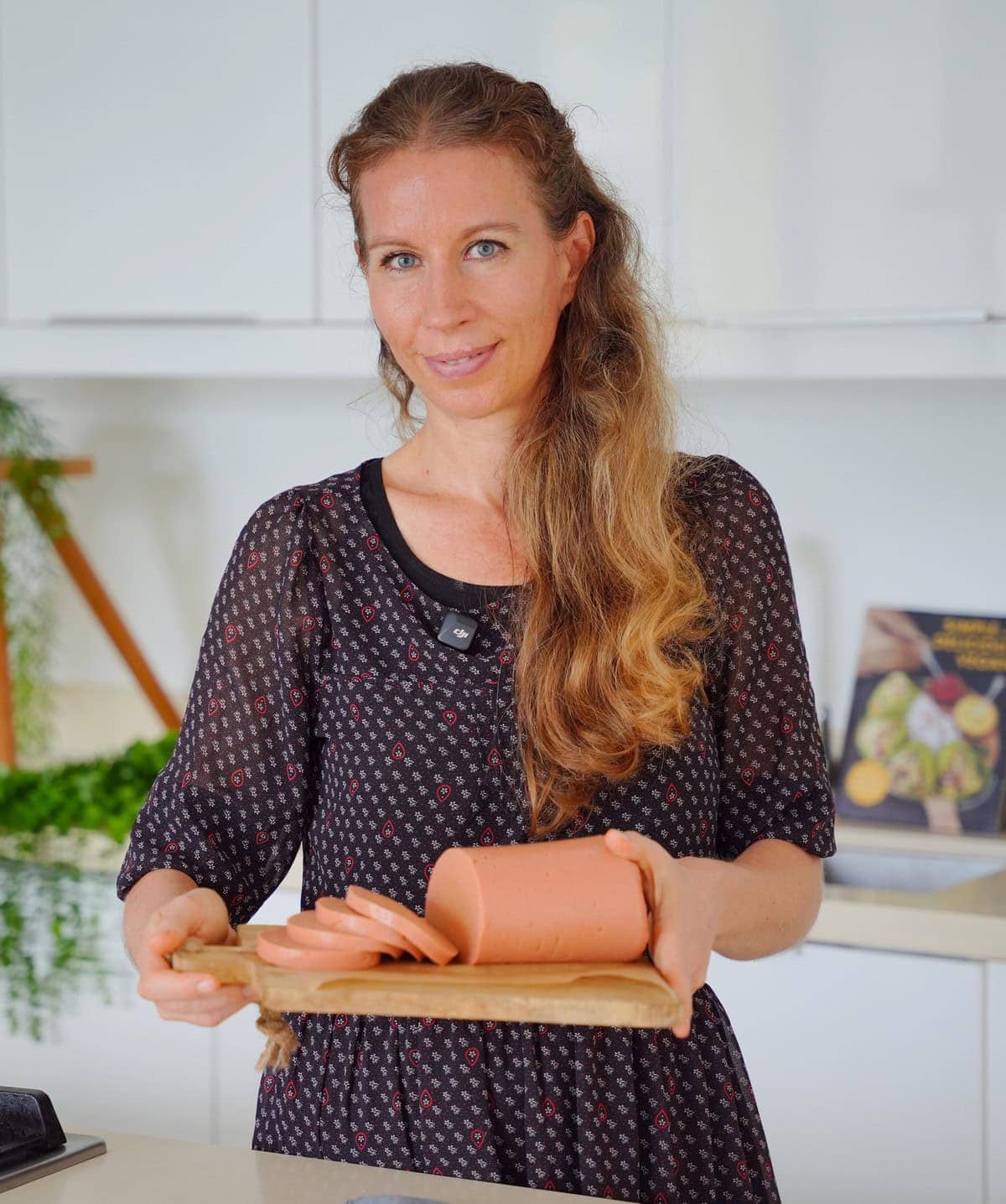 Michaela Vais holds a wooden board with sliced vegan sausage in a bright kitchen. She wears a patterned dress and smiles softly