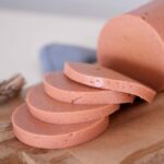 Sliced vegan bologna sausage on a wooden cutting board. Four slices are neatly stacked, showing a smooth texture
