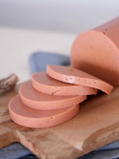 Sliced vegan bologna sausage on a wooden cutting board. Four slices are neatly stacked, showing a smooth texture