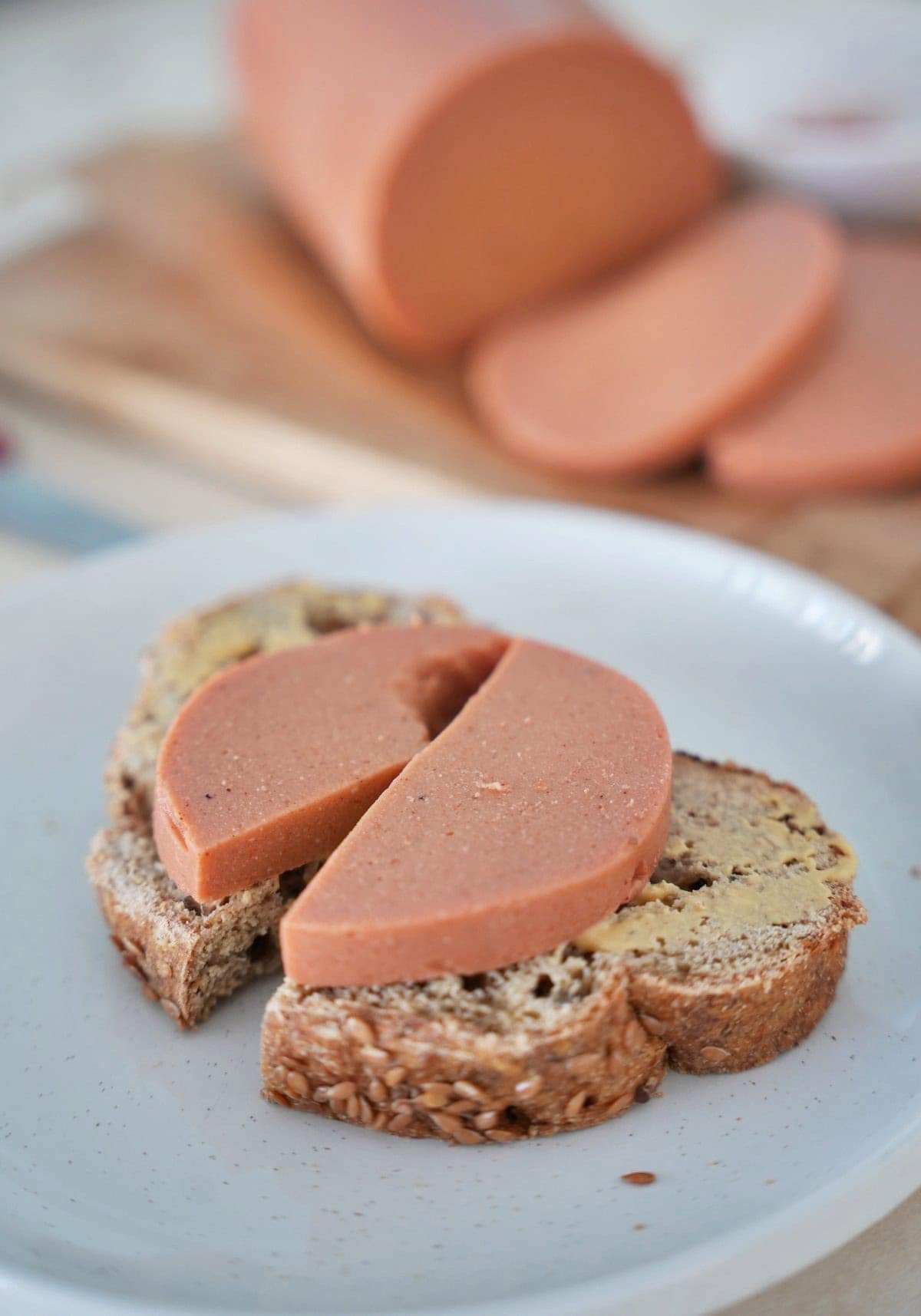 Slices of vegan bologna on whole grain bread, served on a white plate. A whole loaf with slices visible in the background