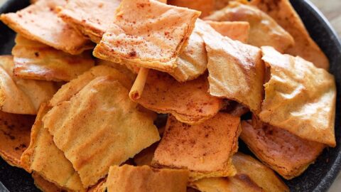 A black bowl filled with crispy, golden-brown rectangular lentil crackers, dusted with paprika
