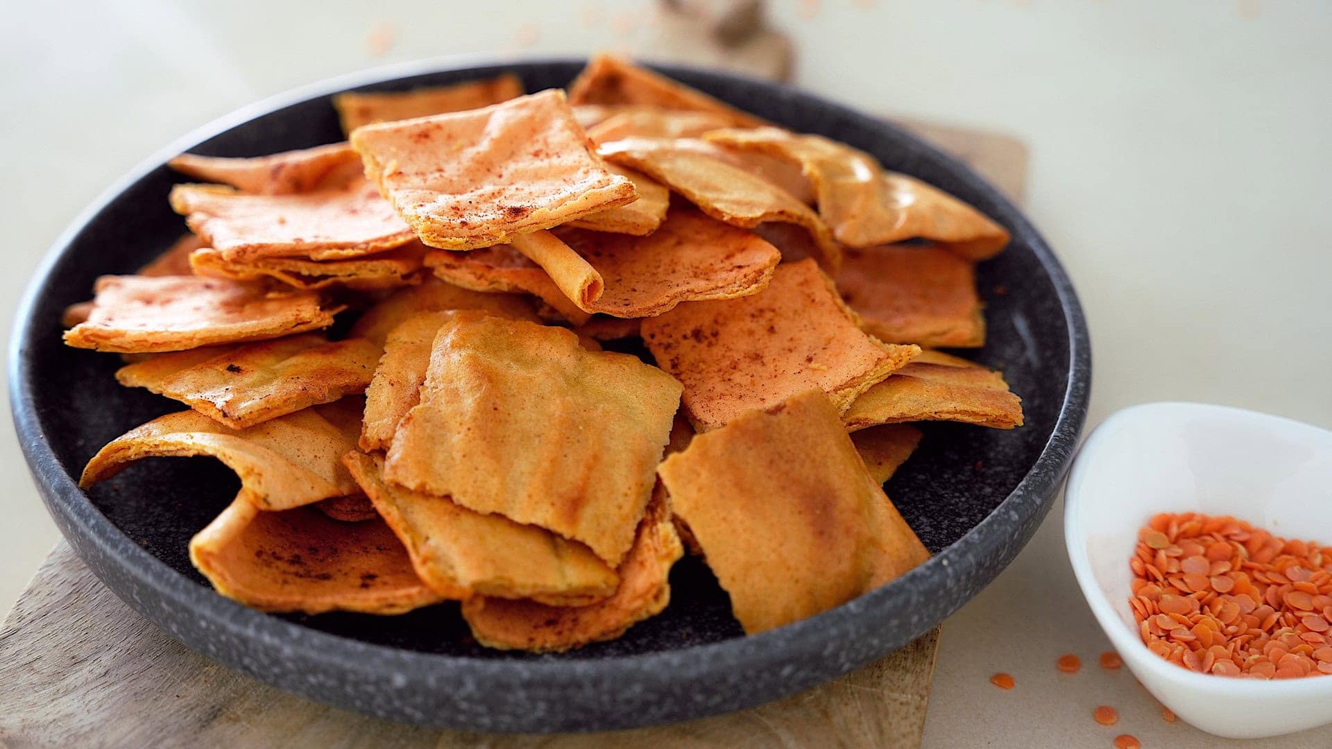 A black plate filled with crispy, golden-brown lentil chips is placed on a wooden board
