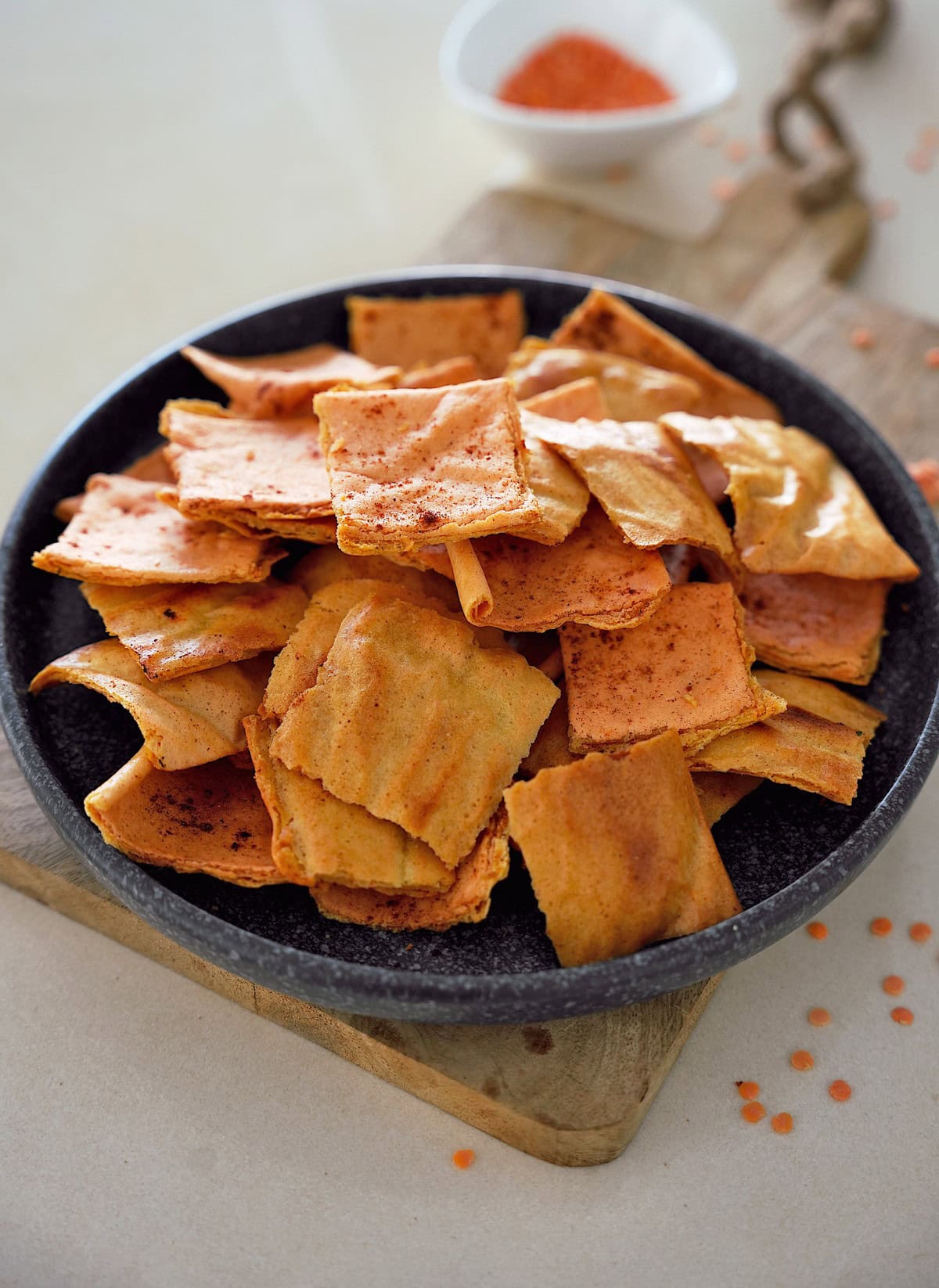 A dark bowl filled with crispy, square, orange lentil crackers is placed on a wooden board