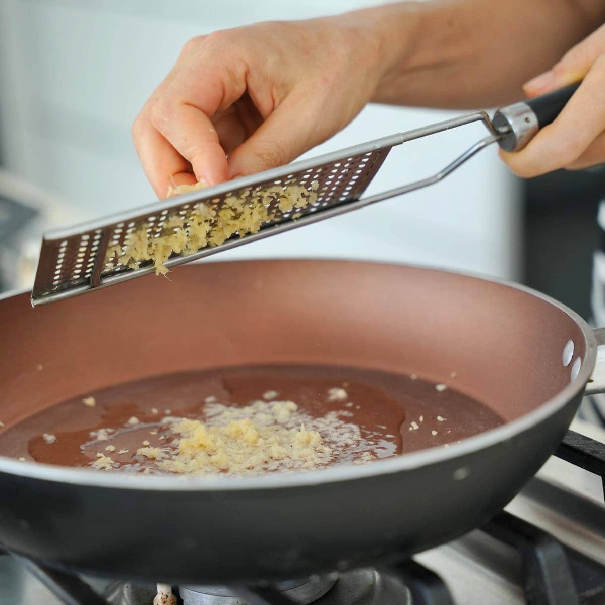 A hand grates fresh ginger over a brown frying pan on a stove