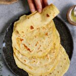 A hand lifts a soft, golden gluten-free tortilla from a stack on a dark plate. Nearby, a small bowl of olive oil