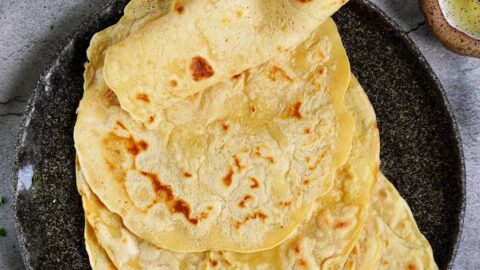 A hand lifts a soft, golden gluten-free tortilla from a stack on a dark plate. Nearby, a small bowl of olive oil
