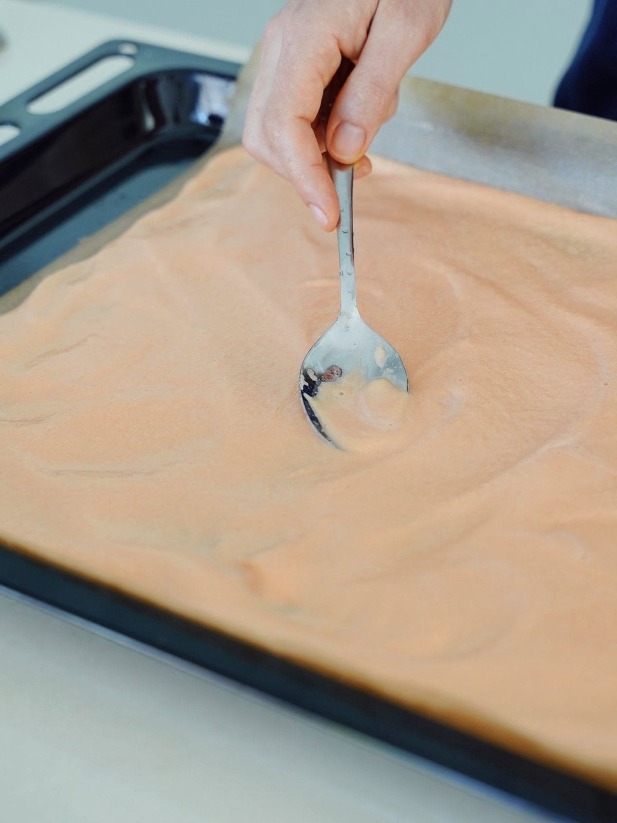 A hand uses a spoon to spread smooth, light orange batter evenly on a baking tray