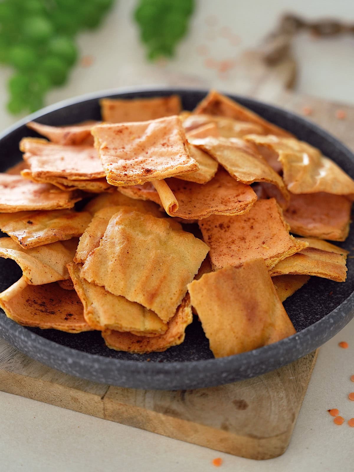 A pile of golden-brown baked lentil crackers is placed on a round, dark stone plate. The chips are slightly curled