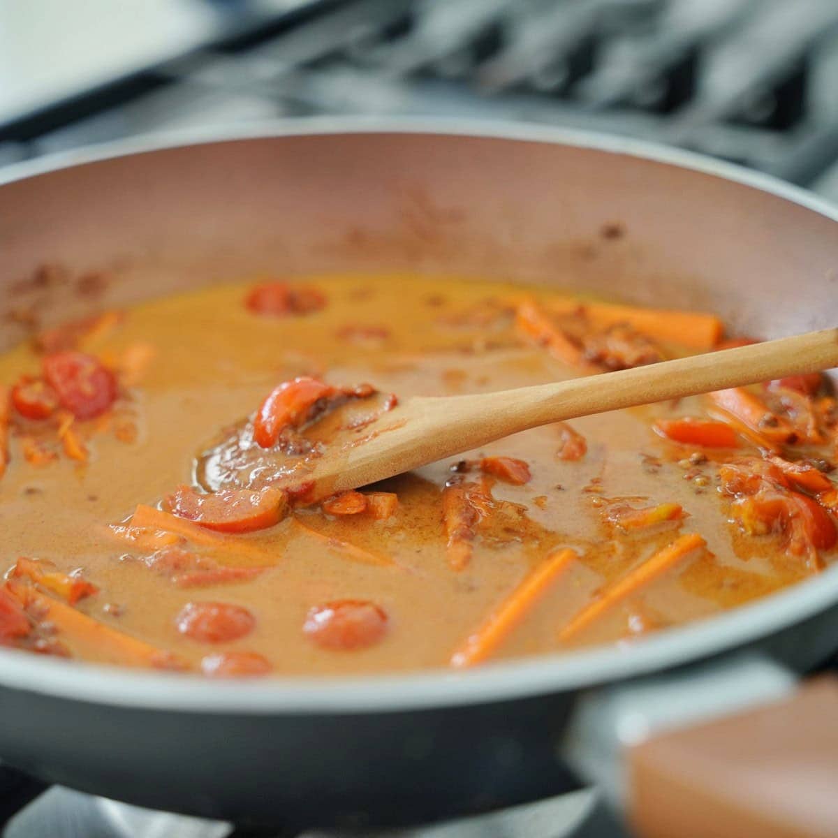 A skillet with curry sauce is simmering on a stovetop, with visible carrot strips and cherry tomatoes