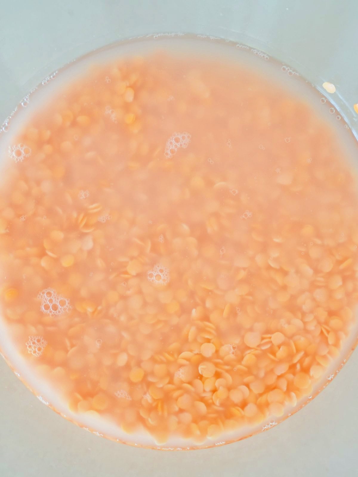 Red lentils submerged in cloudy water within a glass bowl, with small bubbles on the surface