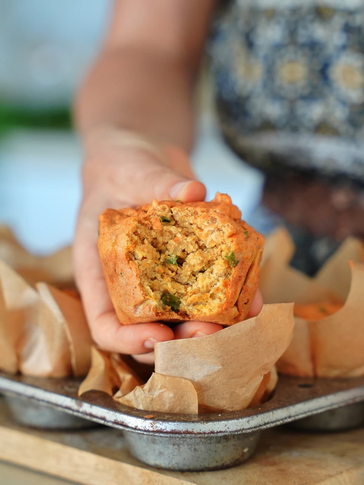 Eine Hand hält einen rustikalen, herzhaften Muffin aus roten Linsen, in dem grüne Kräuterstückchen zu sehen sind