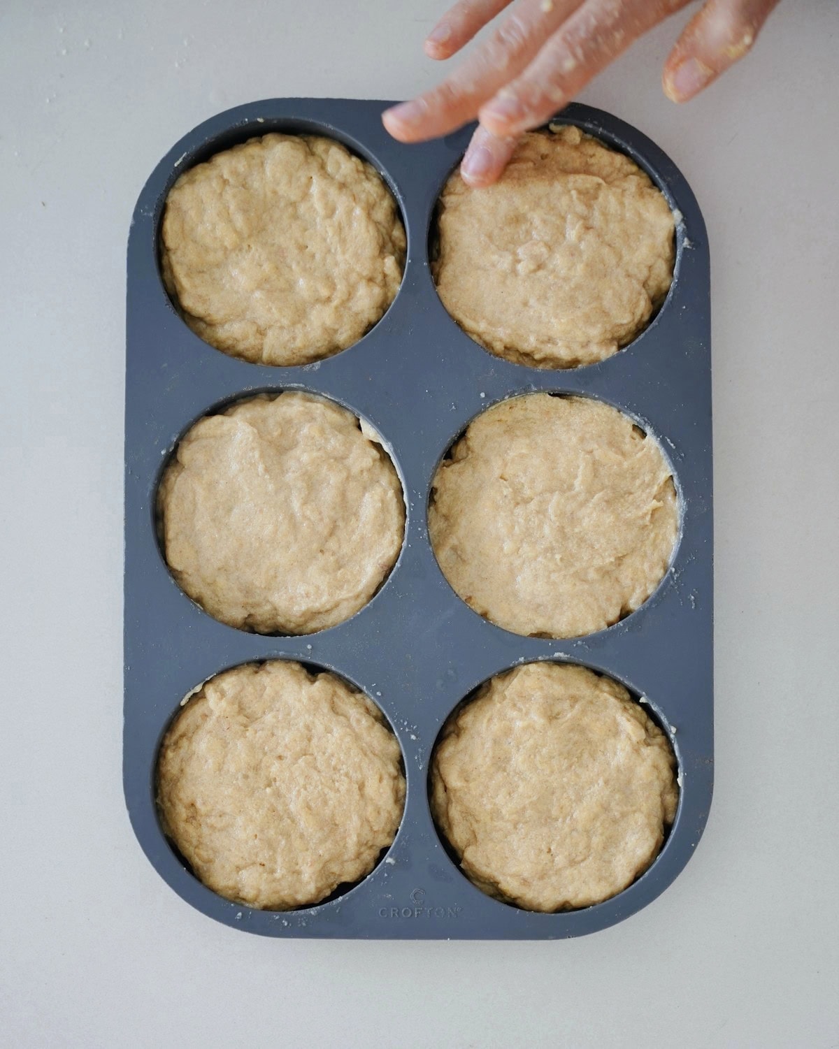 A hand touches a six-cup bun pan filled with raw dough, set on a light surface
