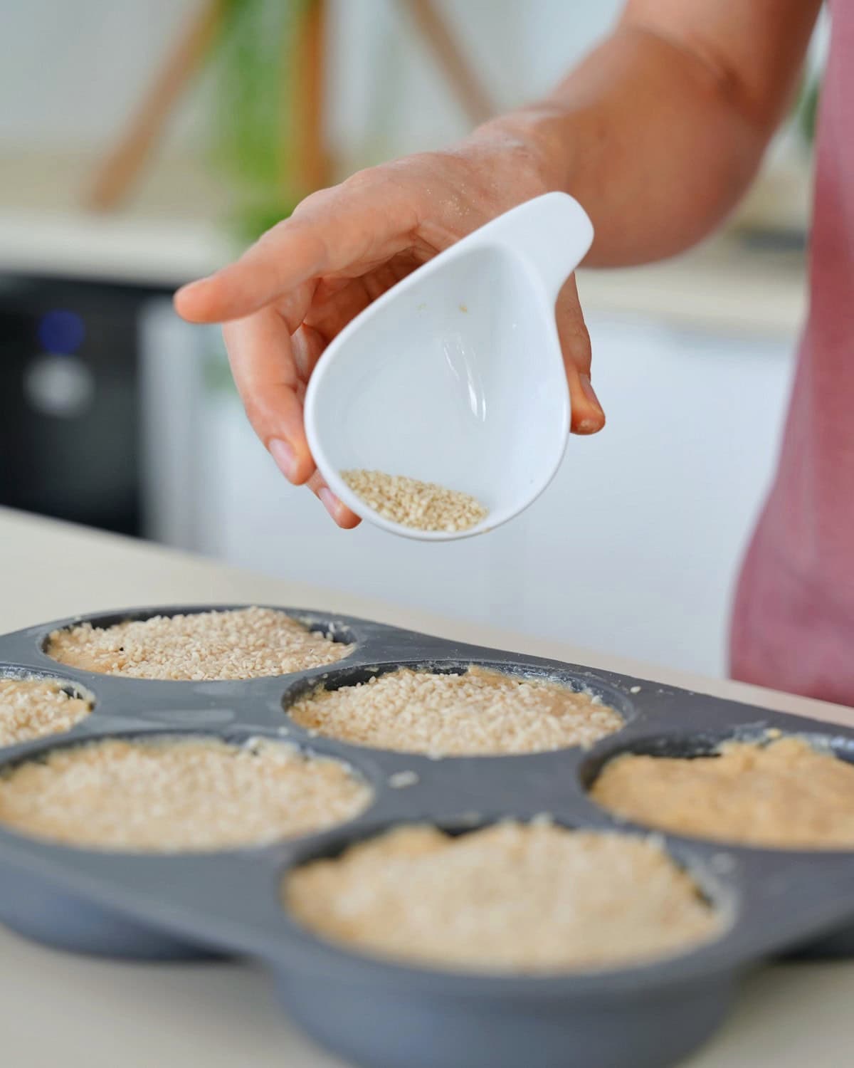 A person sprinkles sesame seeds from a white dish onto bun batter in a gray pan