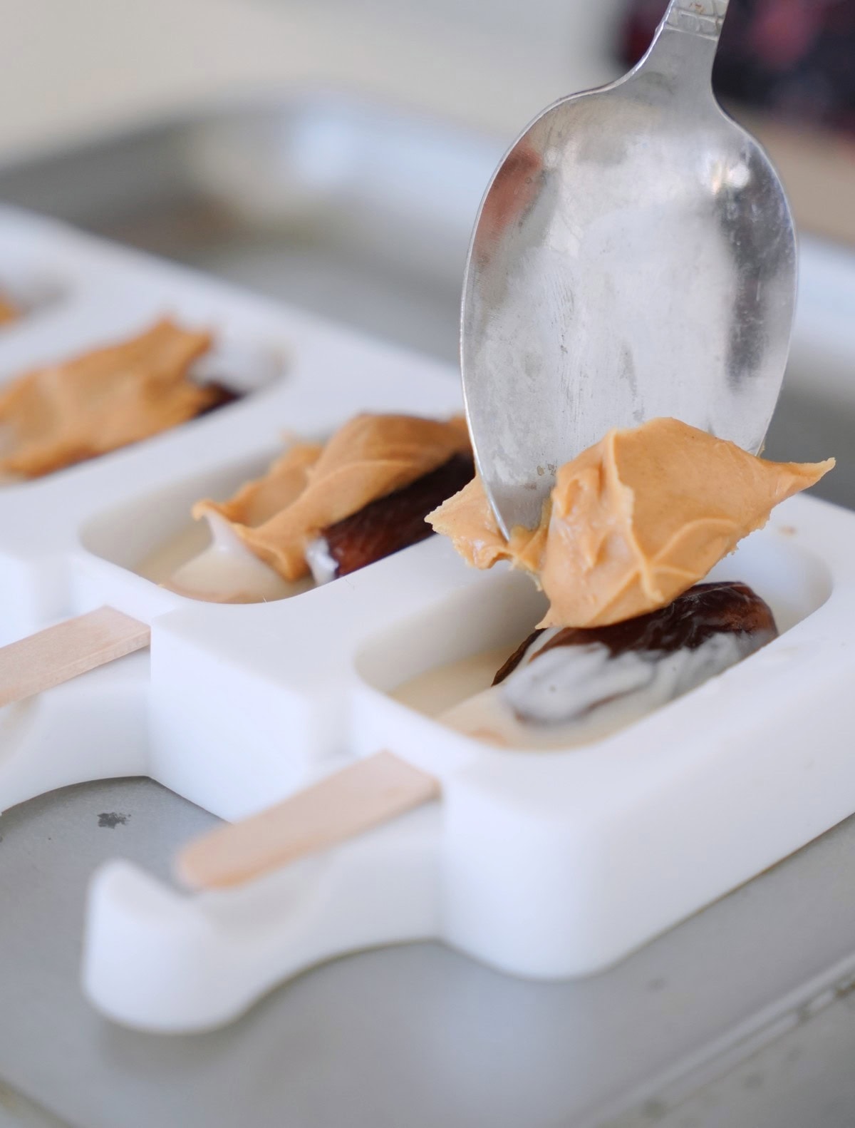 A spoon spreads peanut butter in a white popsicle mold with wooden sticks