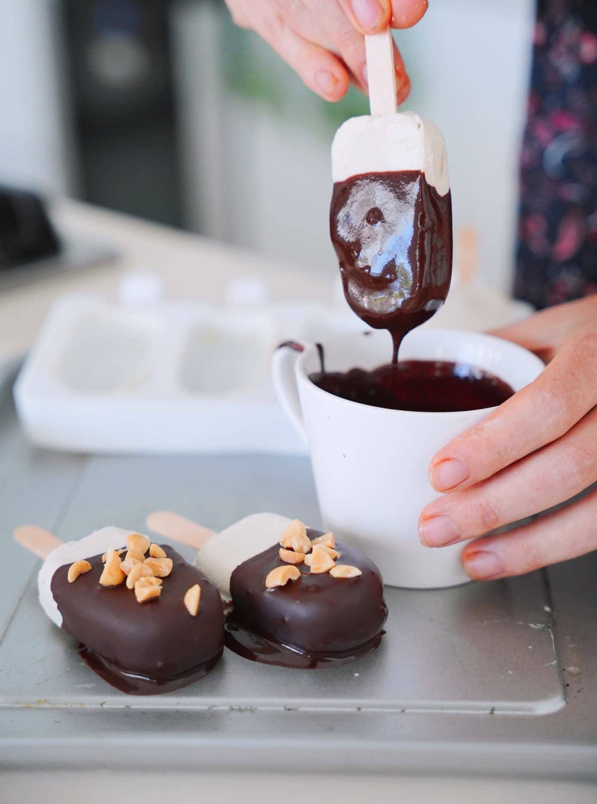 Close-up of a hand dipping a vanilla ice cream bar into melted chocolate. Two finished bars, topped with peanuts, rest on a tray