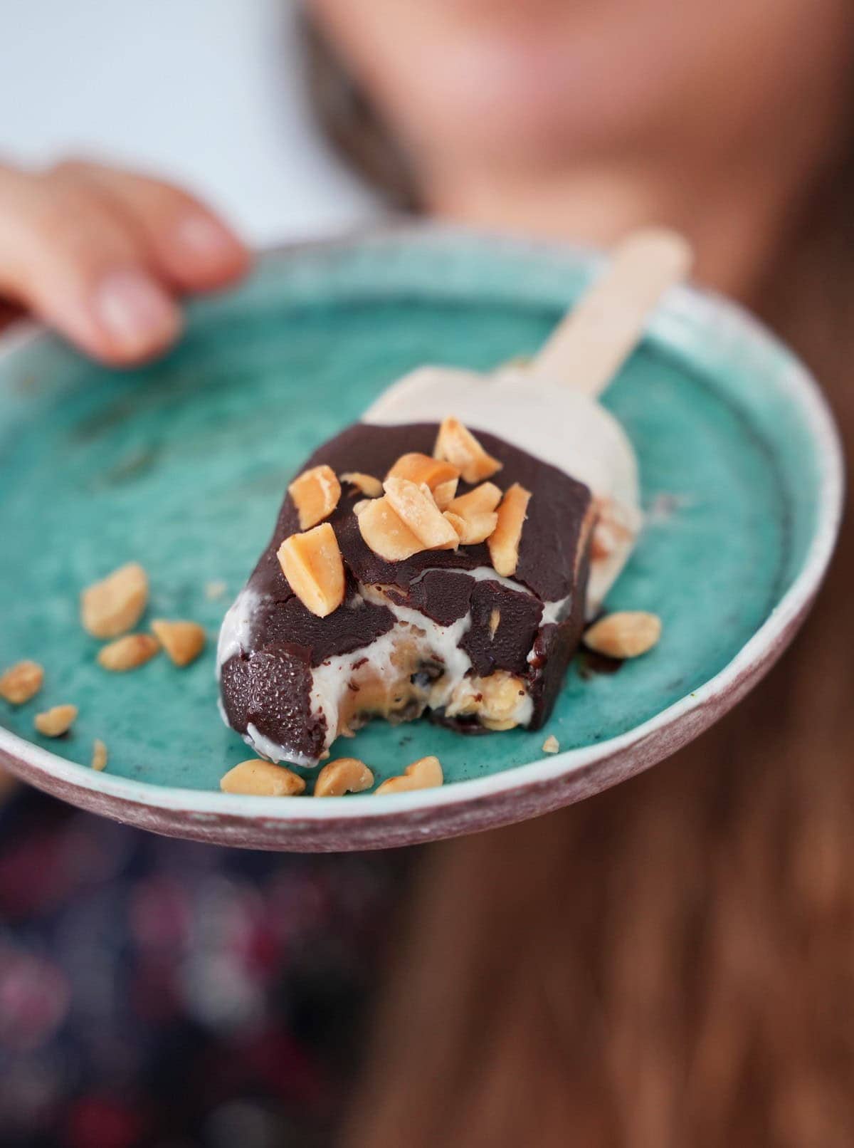 Close-up of a partially eaten chocolate-covered ice cream bar with peanuts on a turquoise plate