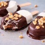 Close-up of three chocolate ice cream bars on sticks, topped with chopped peanuts. One has a bite taken, melting slightly