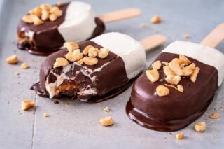 Close-up of three chocolate ice cream bars on sticks, topped with chopped peanuts. One has a bite taken, melting slightly