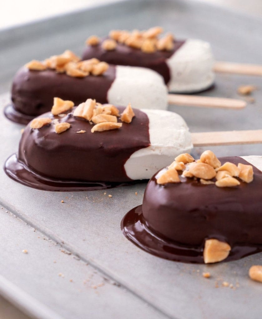 Four white ice cream bars dipped in chocolate, topped with chopped nuts, rest on a metal tray
