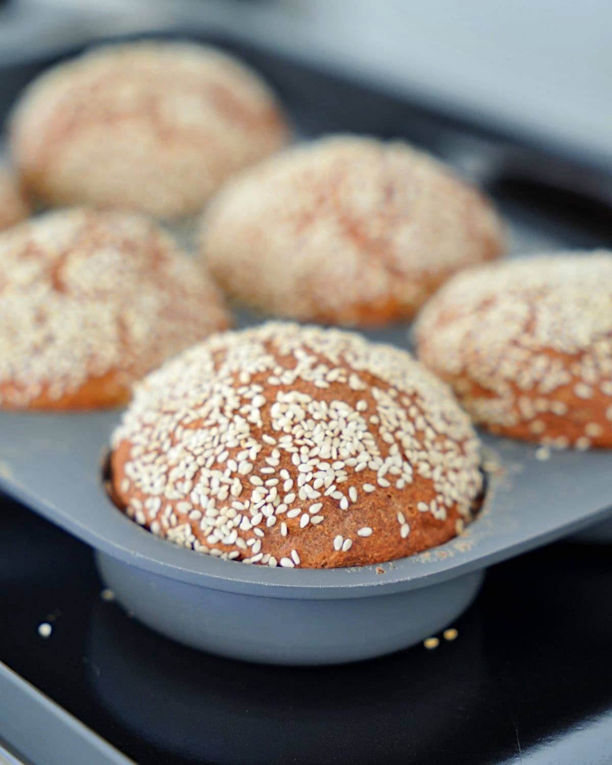 Freshly baked sesame seed-topped gluten-free burger buns sit in a gray tray