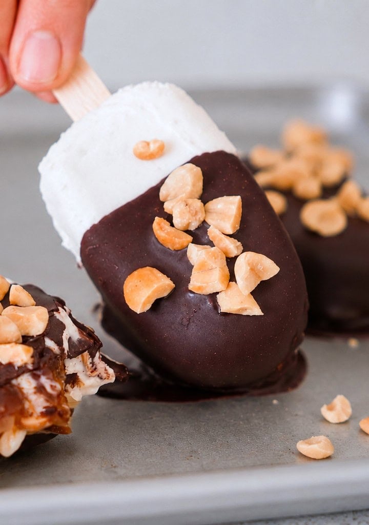 Hand holding a chocolate-dipped ice cream bar with chopped nuts on a tray, alongside two more bars