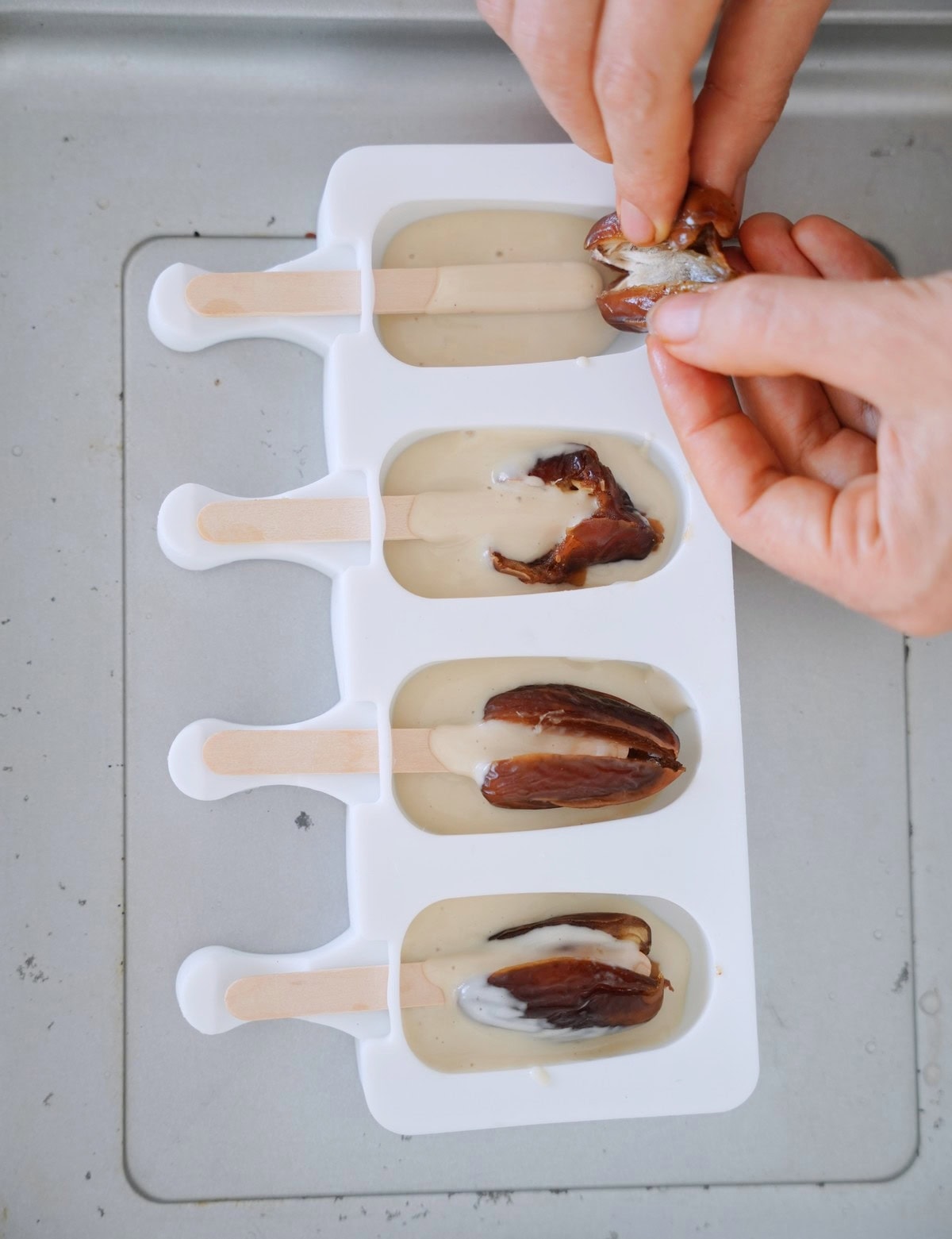 Hands placing a date on a white popsicle mold filled with creamy liquid. Wooden sticks protrude from each mold