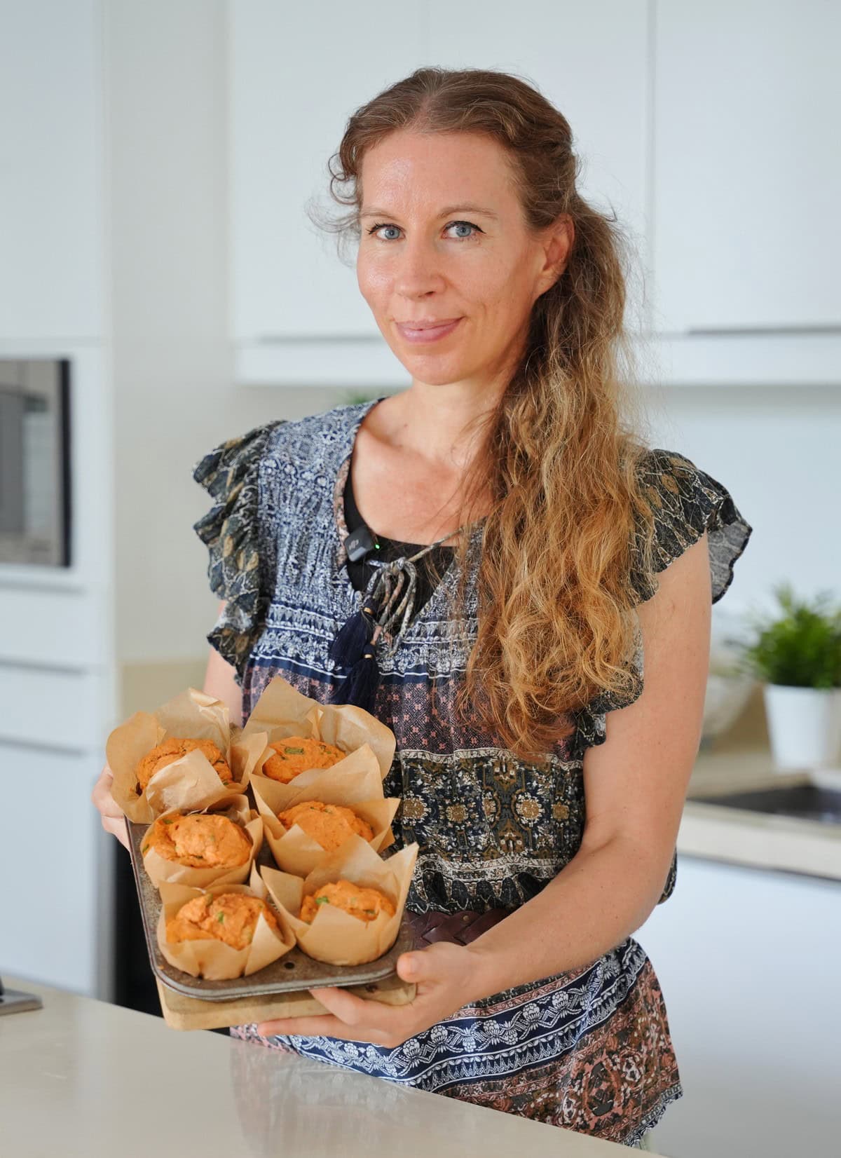 Michaela Vais steht in einer gemusterten Bluse in einer hellen Küche und hält ein Tablett mit frisch gebackenen Linsenmuffins in der Hand