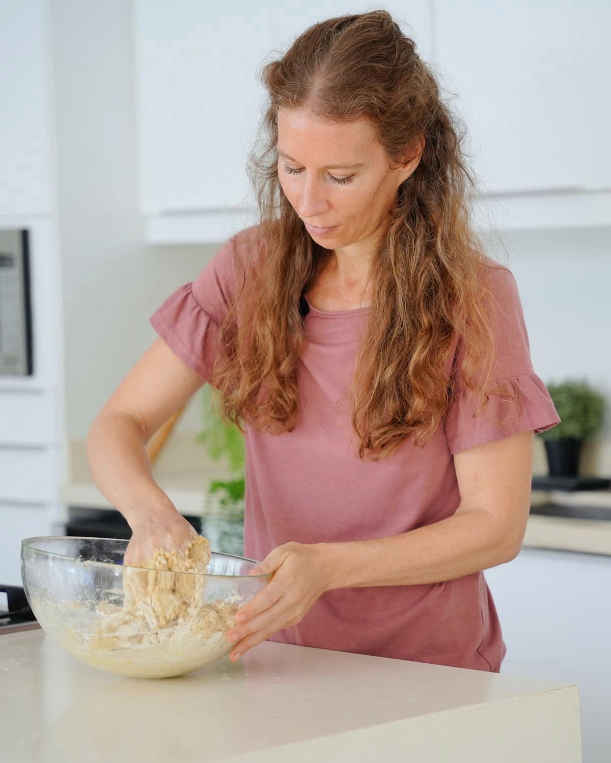 Michaela Vais in a pink shirt kneading dough in a clear bowl, in a bright kitchen