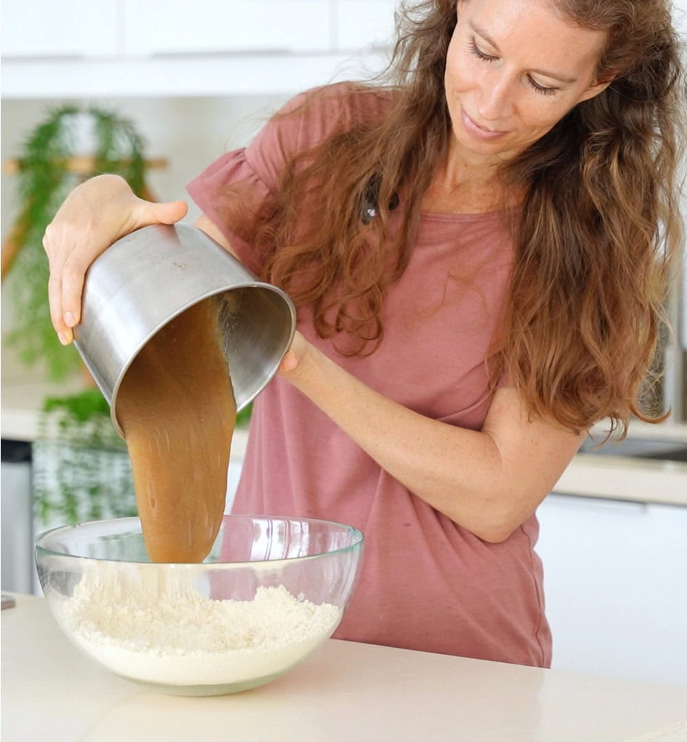 Michaela Vais in a pink shirt pours psyllium gel from a metal jug into a bowl of flour in a bright kitchen