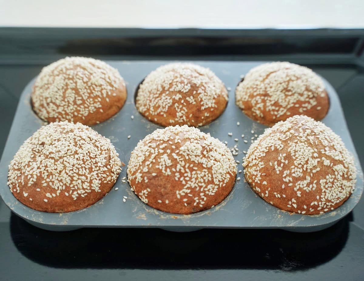Six freshly baked, gluten-free burger buns topped with sesame seeds sit in a gray bun pan