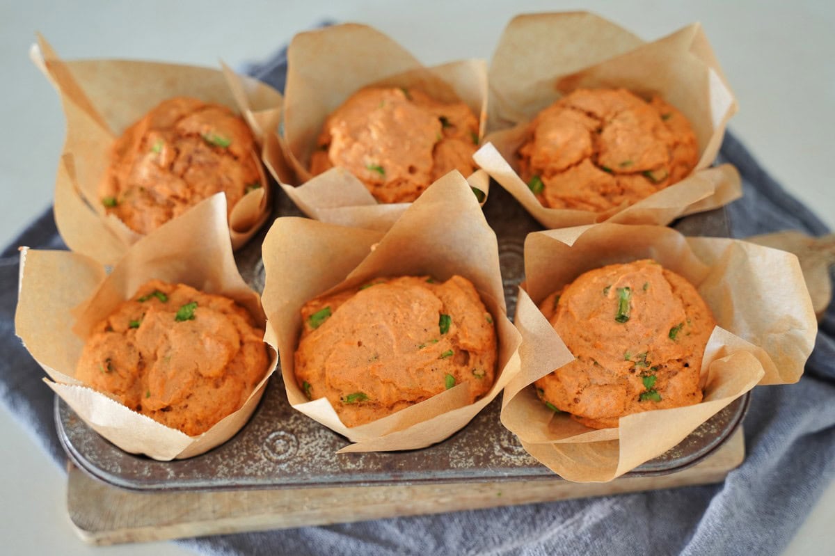 Sechs goldbraune Muffins in Backpapier, garniert mit grünen Kräutern, liegen auf einem Metalltablett