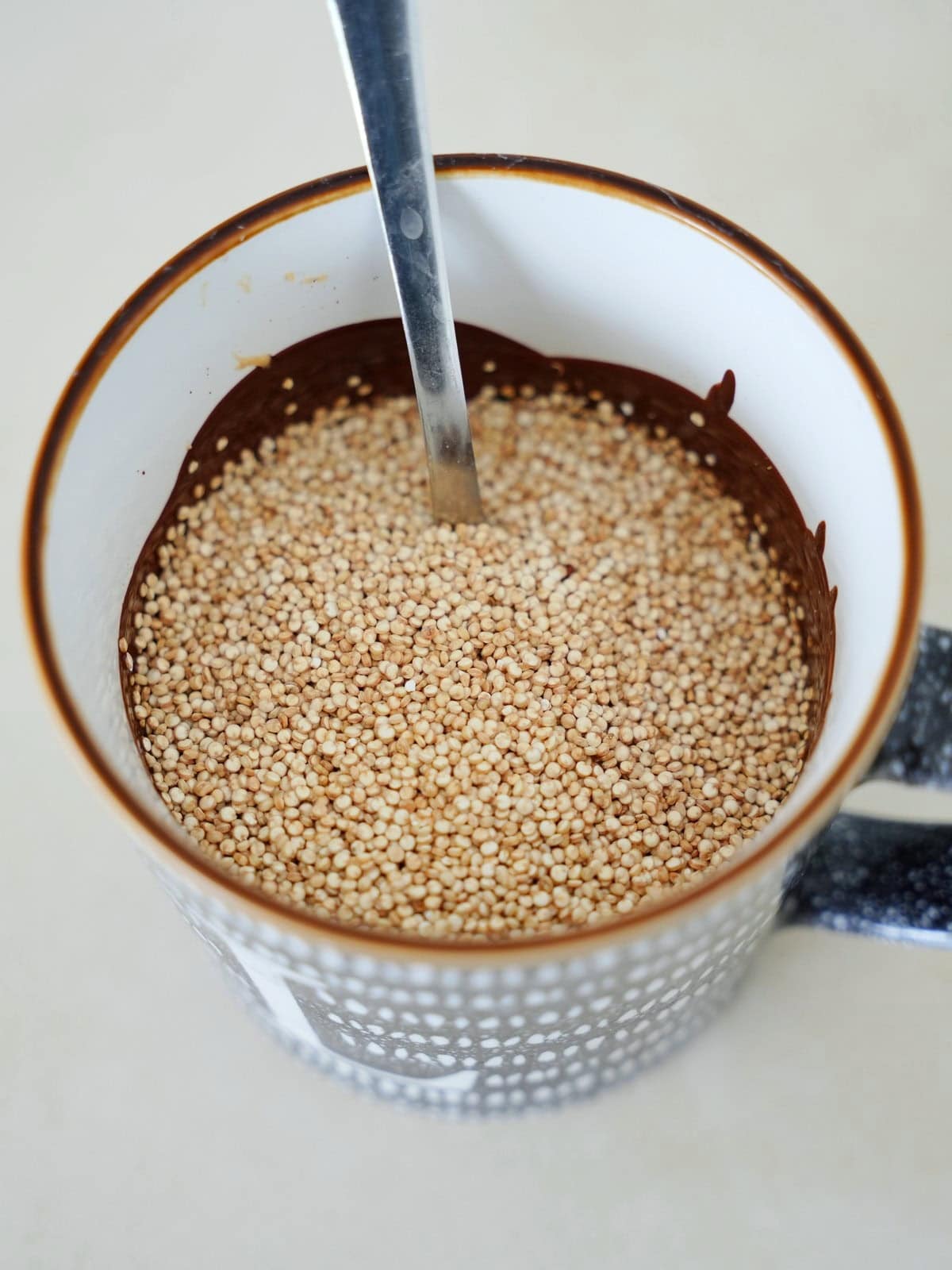 A close-up view of a mug filled with uncooked quinoa seeds and a metal spoon resting inside