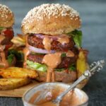 Black bean burger with sesame seed bun, lettuce, onion, and sauce, served with crispy potato wedges on a wooden plate