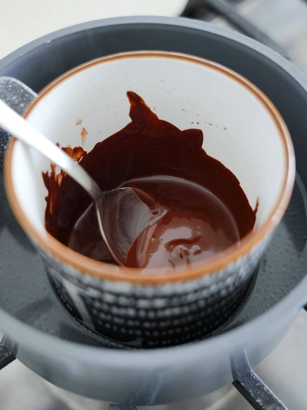 Melted dark chocolate in a mug placed over a simmering pot of water, with a spoon resting inside